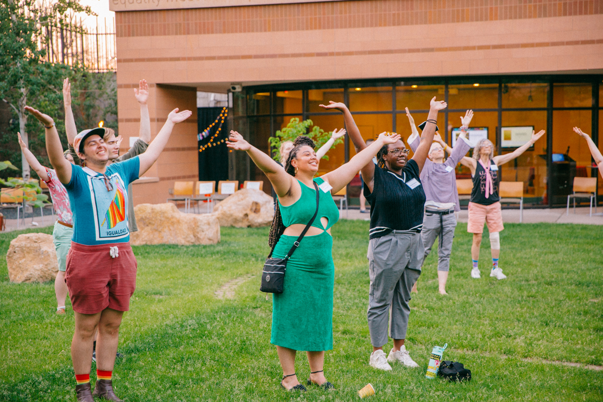 Storytelling participants are spread out standing in a green space, arms open wide above their heads.