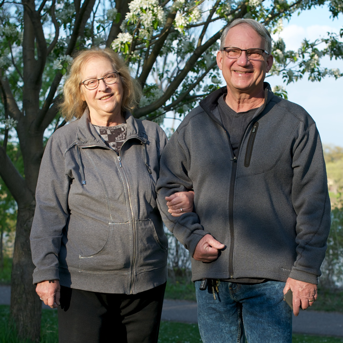 A couple stands arm in arm in a park, smiling at the camera.