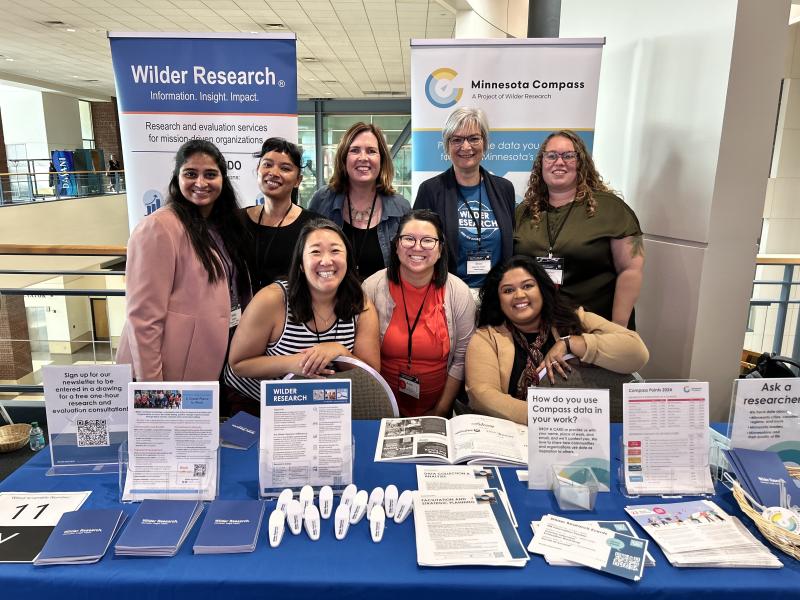 A group of eight women post behind a Wilder Research exhibit table. Three women are seated and five additional women are standing behind them. Handouts and giveaways are spread across the table in the foreground.