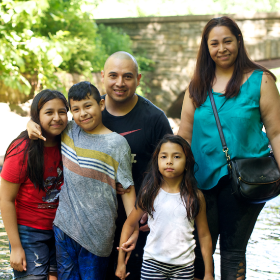 A family of two adults and three children wades in a stream in a park setting. 