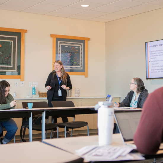 A standing woman leads a discussion with people seated around a conference room table.