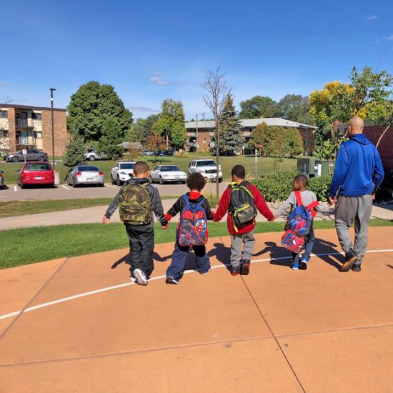 Four children and one adult hold hands and walk away from the camera with a school setting in the background