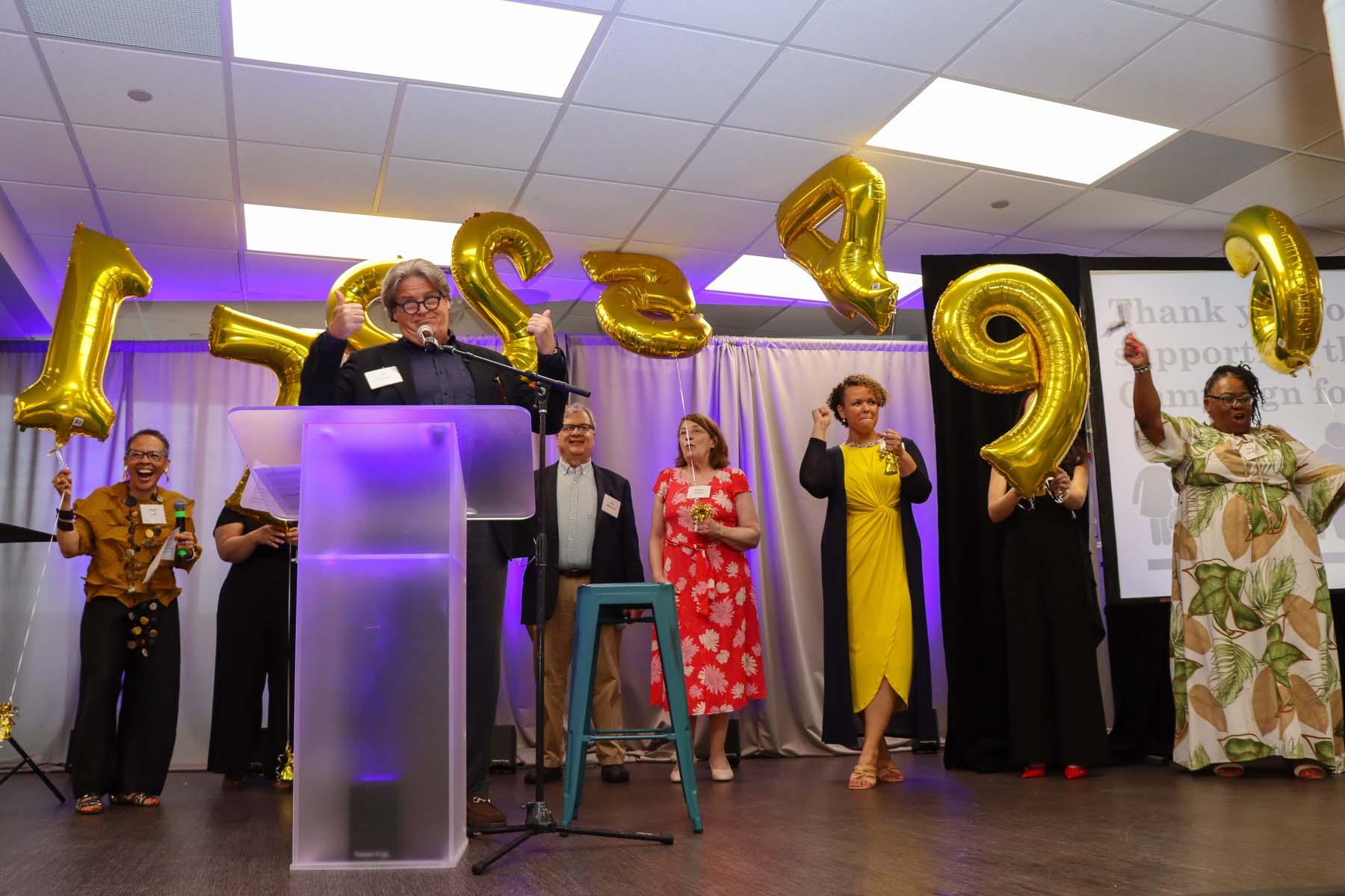 A group of people stand together holding helium balloons in the shape of numbers