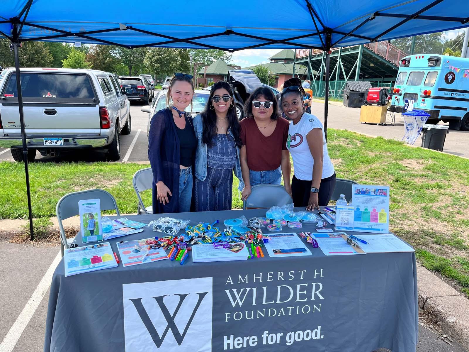 four women stand behind a table with a Wilder table cloth and supplies