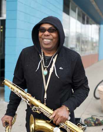 An adult stands on the sidewalk in front of a brick building, smiling and holding a saxophone.