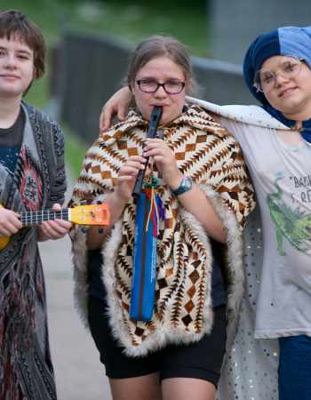 Three young people holding musical instruments stand together on a sidewalk.